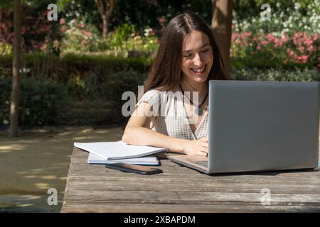 Una giovane studentessa del college a un tavolo del campus sorride mentre partecipa a una riunione virtuale con il suo laptop. Inizio delle lezioni, vita studentesca, technolo Foto Stock