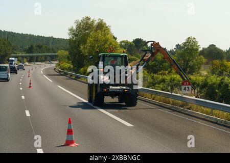 Salles-d'Aude, Francia - 16 maggio 2023: Un addetto alla manutenzione gestisce un escavatore sul lato di un'autostrada, con coni stradali impostati per la sicurezza durante Foto Stock