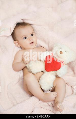 Un bambino sdraiato su una coperta rosa che regge un orsacchiotto bianco con un cuore rosso Foto Stock