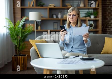 Una donna matura si siede sul divano con un laptop e uno smartphone, gestendo le finanze e rivedendo le bollette in un accogliente salotto. Foto Stock