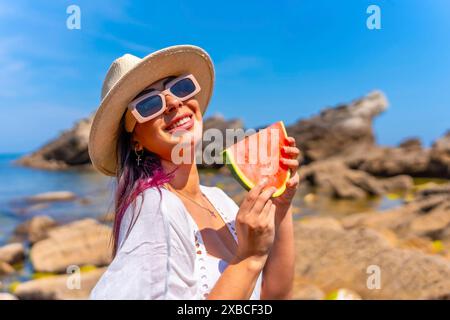 Una donna tiene in mano una fetta di anguria mentre indossa un cappello di paglia e occhiali da sole. Lei sorride e si sta godendo la giornata di sole Foto Stock