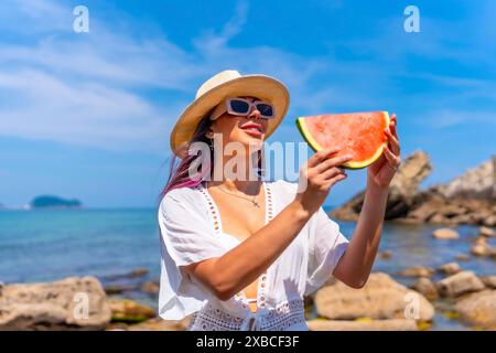 Una donna tiene in mano una fetta di anguria mentre si trova in spiaggia. La scena è luminosa e soleggiata, con l'oceano sullo sfondo. Il Foto Stock