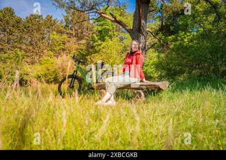 Una donna siede su una panchina nella foresta, accanto a lei una bicicletta, circondata da un prato soleggiato, e-Bike, Foresta Nera, Gechingen, Germania Foto Stock