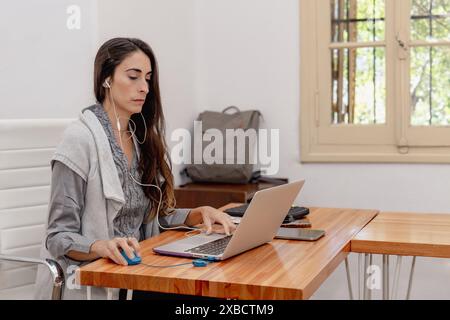 Una donna latina di mezza età, dai capelli lunghi, siede sulla scrivania dell'ufficio di casa, concentrandosi sul laptop mentre indossa le cuffie. Foto Stock