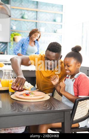 Giovane uomo e ragazza afro-americana che pranzano insieme a tavola. Donna afroamericana che prepara il cibo in una cucina moderna con piastrelle blu Foto Stock