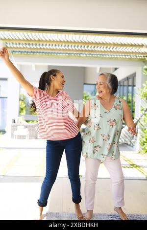 Nonna asiatica e nipote birazziale ballano gioiosamente insieme a casa. Soggiorno luminoso e arioso con grandi finestre e arredi moderni, creat Foto Stock