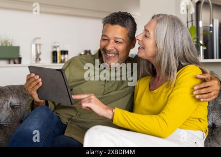 Una coppia birazziale matura si siede sul divano, sorride e guarda un tablet, a casa. Si trovano in un intimo soggiorno con decor moderno e alcune piante Foto Stock