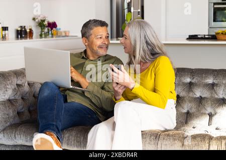 Una coppia birazziale matura si siede su un divano, utilizzando un computer portatile e uno smartphone, a casa. Sono in un moderno e luminoso soggiorno con cucina in backgro Foto Stock