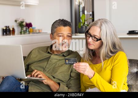 Una coppia matura birazziale usa un laptop e una carta di credito insieme in una cucina moderna, a casa Foto Stock