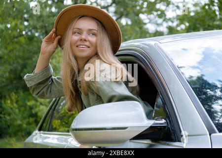 Donna bionda con cappello che sporge la testa dalla vettura del parabrezza. I giovani turisti esplorano i viaggi locali, trasformandoli in momenti autentici. Emozioni vere espressioni di fuga e di rinfrescarsi rilassarsi all'aria aperta e pulita Foto Stock