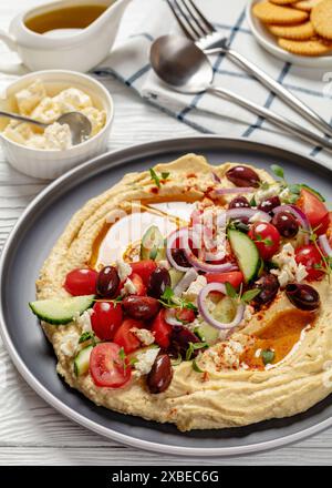 Hummus greco carico di cetrioli freschi, olive, pomodori, feta, cipolle rosse e timo su un tavolo di legno bianco con cracker, V verticale Foto Stock