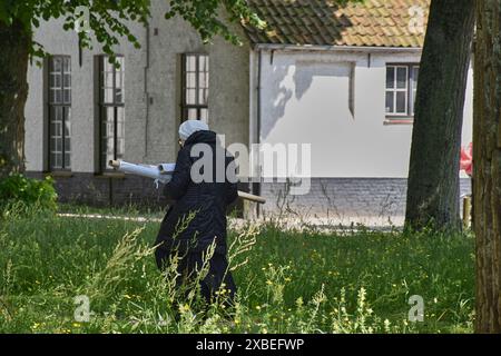 Bruges, Bruxelles; giugno 2024; una vecchia suora cammina attraverso il giardino del convento. Caricato con materiale laminato. Berretto bianco e nero Foto Stock