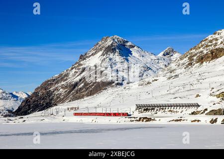 Berina, Svizzera - gennaio 06. 2023: La ferrovia retica rossa passa lungo il lago bianco ghiacciato con la cima delle Alpi Piz Lagalb al backgroun Foto Stock