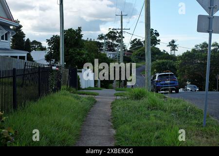 Vista in cima alla collina di via Firenze, carina, sentiero in cemento verde, erboso e coperto, pali elettrici, auto parcheggiata, nel tardo pomeriggio a Brisbane Foto Stock