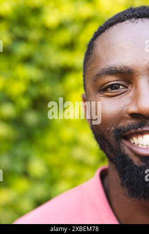 Giovane afroamericano sorridente calorosamente, indossa una camicia rosa, a casa. Il verde rigoglioso crea uno sfondo verde brillante e naturale, che si aggiunge a liv Foto Stock