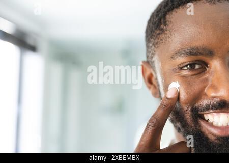 Giovane afroamericano che applica crema facciale, sorridendo calorosamente, a casa. Bagno luminoso con finiture moderne sullo sfondo, piastrelle bianche e grande Foto Stock