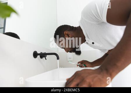 Giovane afroamericano che si lava i denti in un bagno moderno, a casa. Decorazioni bianche minimaliste con eleganti finiture nere che creano uno stile elegante e pulito Foto Stock
