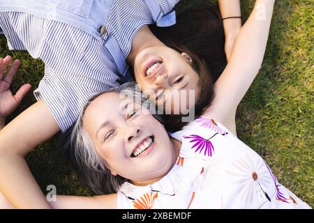 Nonna asiatica e nipote birazziale sdraiati sull'erba sorridendo insieme. Godetevi una giornata di sole in un giardino, circondato da vegetazione lussureggiante e vivace Foto Stock
