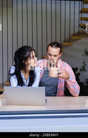 Giovani coppie diverse sono sedute insieme, utilizzando un notebook. Sono in una casa moderna con una scala e piante sullo sfondo, inalterate Foto Stock