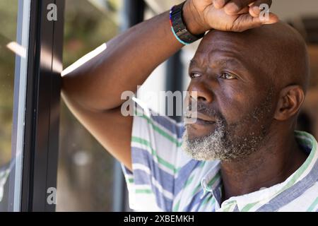 Un uomo afroamericano maturo che guarda attentamente fuori dalla finestra. La luce del sole filtra attraverso il vetro, illuminando la sua espressione contemplativa in un'atmosfera accogliente Foto Stock