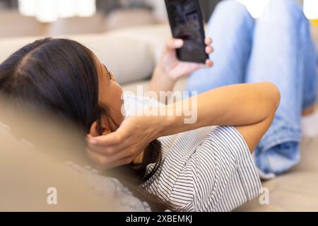 Giovane donna birazziale che si rilassa sul divano guardando lo smartphone, a casa. Soggiorno accogliente con illuminazione soffusa e arredi confortevoli che creano calore e. Foto Stock