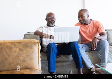 Padre e figlio afroamericani stanno guardando un portatile insieme su un divano. Si trovano in un accogliente soggiorno con un divano beige e un tavolino da caffè in vetro, Foto Stock