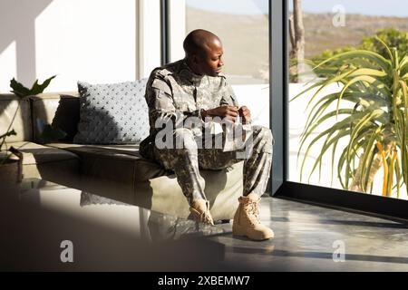 Giovane soldato afroamericano in uniforme militare seduto sul divano a guardare una lettera. La luce del sole scorre attraverso grandi finestre, illuminando in modo moderno Foto Stock