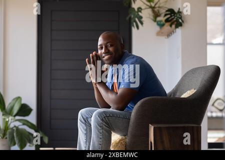 Giovane afroamericano seduto sulla sedia, sorridente e rilassato. Interni moderni con piante e una porta di sfondo scuro che creano un intimo Foto Stock