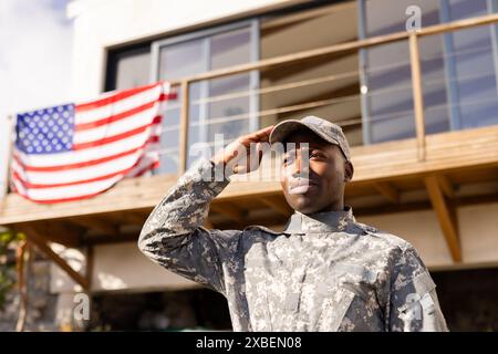 Giovane soldato afroamericano in uniforme militare che saluta. In piedi davanti alla casa con bandiera americana appesa sul balcone, mostrando orgoglio e sollievo Foto Stock