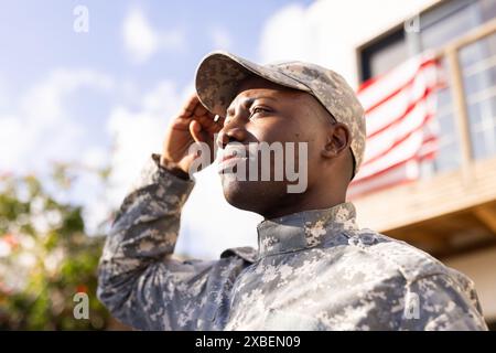 Giovane soldato afroamericano in uniforme militare che saluta. Bandiera americana e casa sullo sfondo, inalterata Foto Stock