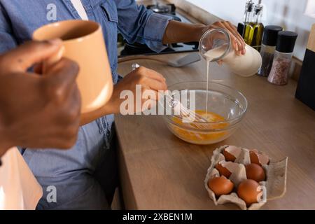 Una coppia di persone diverse prepara la colazione, sbattendo le uova e aggiungendo il latte in un recipiente. Sono in una cucina moderna con vari ingredienti di cottura Foto Stock