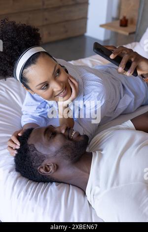 Giovane coppia birazziale che si rilassa sul letto e sorride sullo smartphone. Accogliente camera da letto con mobili in legno e illuminazione soffusa crea un'atmosfera calda e intima. Foto Stock