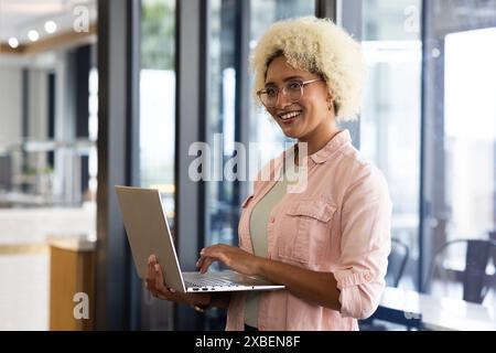 Giovane donna birazziale che tiene un notebook e sorride con fiducia in ufficio. Spazio di lavoro moderno con pareti divisorie in vetro e luce naturale, Foto Stock