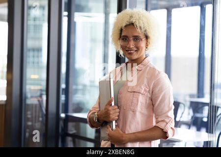 Giovane donna birazziale che tiene un laptop, sorride alla macchina fotografica in ufficio. Pareti in vetro e mobili moderni che creano un backgroun professionale ed elegante Foto Stock