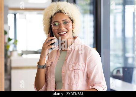 Giovane donna birazziale con gli occhiali sorridenti mentre è su smartphone in ufficio . Pareti in vetro e decor moderno che creano uno sfondo professionale e moderno Foto Stock