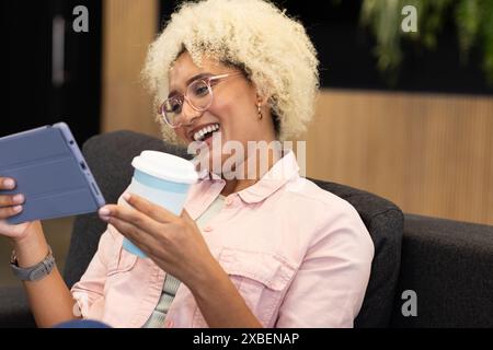 Giovane donna birazziale in ufficio che tiene un tablet e una tazza di caffè, sorridente. Sedersi sul divano nella moderna area salotto dell'ufficio con pannelli in legno Foto Stock