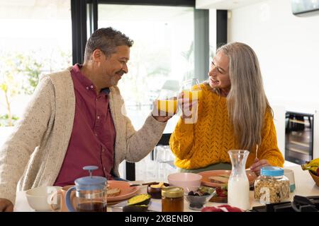Una coppia birazziale matura sorridente e tostata con succo di frutta a casa, a casa. Cucina luminosa con elettrodomestici moderni e una varietà di colazioni Foto Stock