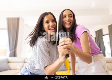 Due giovani sorelle birazziali si divertono insieme con il karaoke, sorridendo e tenendo il microfono, a casa. Sono in un luminoso e moderno soggiorno con grande vittoria Foto Stock