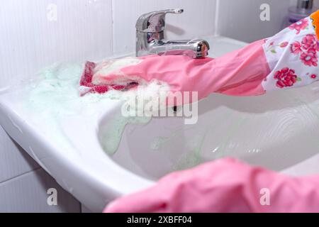 Donna con guanti rosa che pulisce lavabo in ceramica bianca con spugna e detergente, primo piano Foto Stock