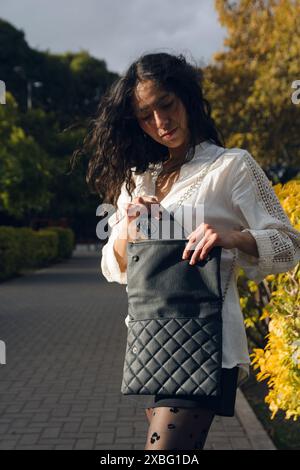 Una donna latina che prende il telefono dalla sua borsa sulla piazza. Indossa una camicia bianca e calze nere, immagine verticale, social media e techno Foto Stock