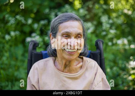 Vecchia donna indiana (Asia meridionale) di 90 anni sorridente, indossa abiti casual seduti su una sedia a rotelle all'aperto in natura, Regno Unito Foto Stock