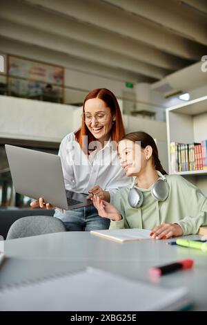 Redhead Woman fa da tutor a una ragazza adolescente in una biblioteca, entrambe focalizzate su un portatile, impegnata in lezioni dopo la scuola. Foto Stock