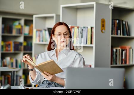 Una donna rossa è profondamente coinvolta nella lettura di un libro all'interno di una biblioteca, circondata da scaffali di libri. Foto Stock