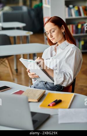 Una tutor donna con la testa rossa siede alla sua scrivania, concentrandosi sulla scrittura su un notebook dopo una lezione online. Foto Stock