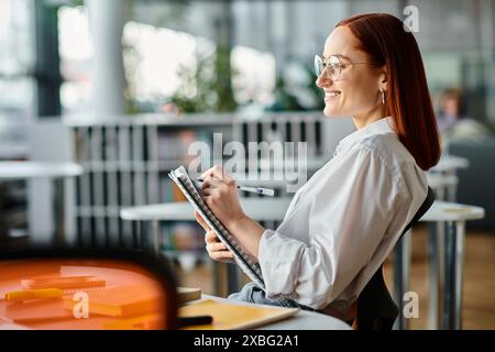 Una donna dai capelli rossi si siede a una scrivania, scrive su una clipboard mentre impartisce una lezione online usando un laptop dopo la scuola. Foto Stock