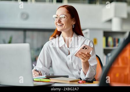 Una tutor rossa insegna online dopo la scuola, seduto a una scrivania con un portatile e un cellulare. Foto Stock