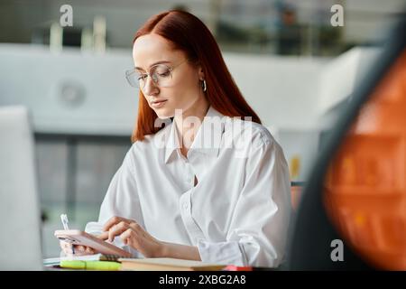 Una tutor rossa si occupa di insegnamento online, seduto a una scrivania e interagire con uno smartphone dopo l'orario di scuola. Foto Stock