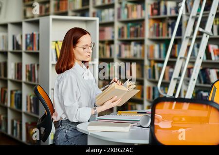 Una donna con la testa rossa siede a un tavolo in una biblioteca, coinvolta nella lettura di un libro. Foto Stock