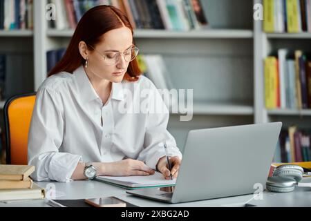 Una tutor rossa si concentra sull'insegnamento di una lezione dopo la scuola mentre lavora su un notebook in una biblioteca. Foto Stock