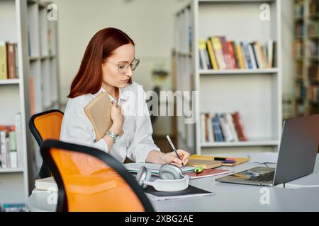 Una tutor donna con i capelli rossi che fornisce una lezione di after-School online, utilizzando un portatile a una scrivania in un ambiente libreria. Foto Stock